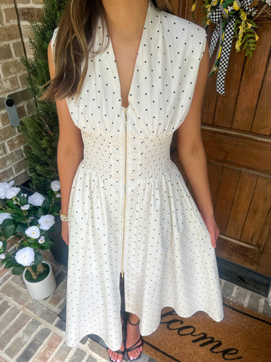 Woman wearing a white polka dot dress standing on a porch with plants and a welcome mat.