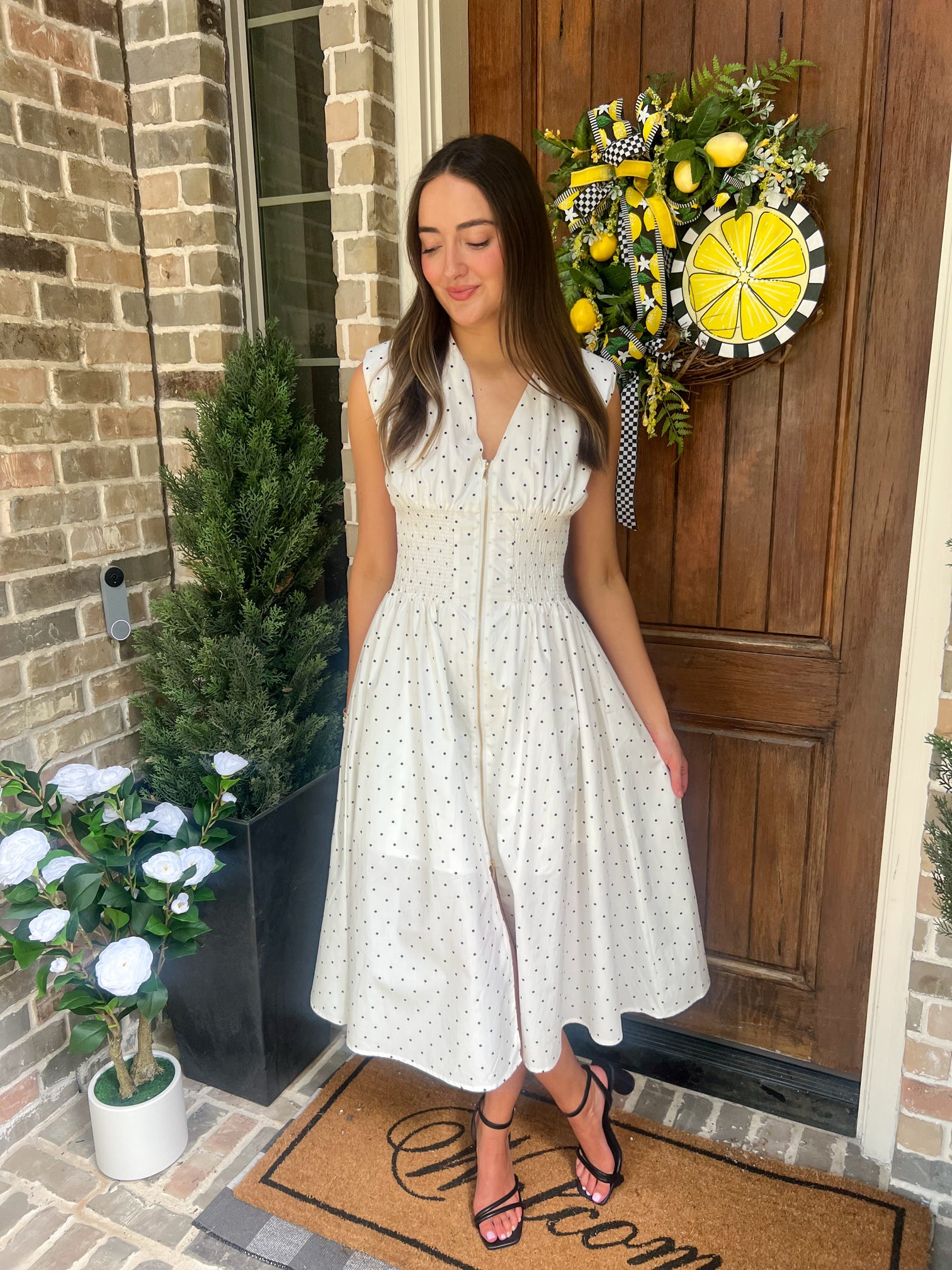 Woman in a white with small black polka dots dress standing on a porch with decorative elements.