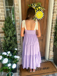 Woman in a lavender dress standing in front of a door with a wreath, surrounded by plants and flowers.