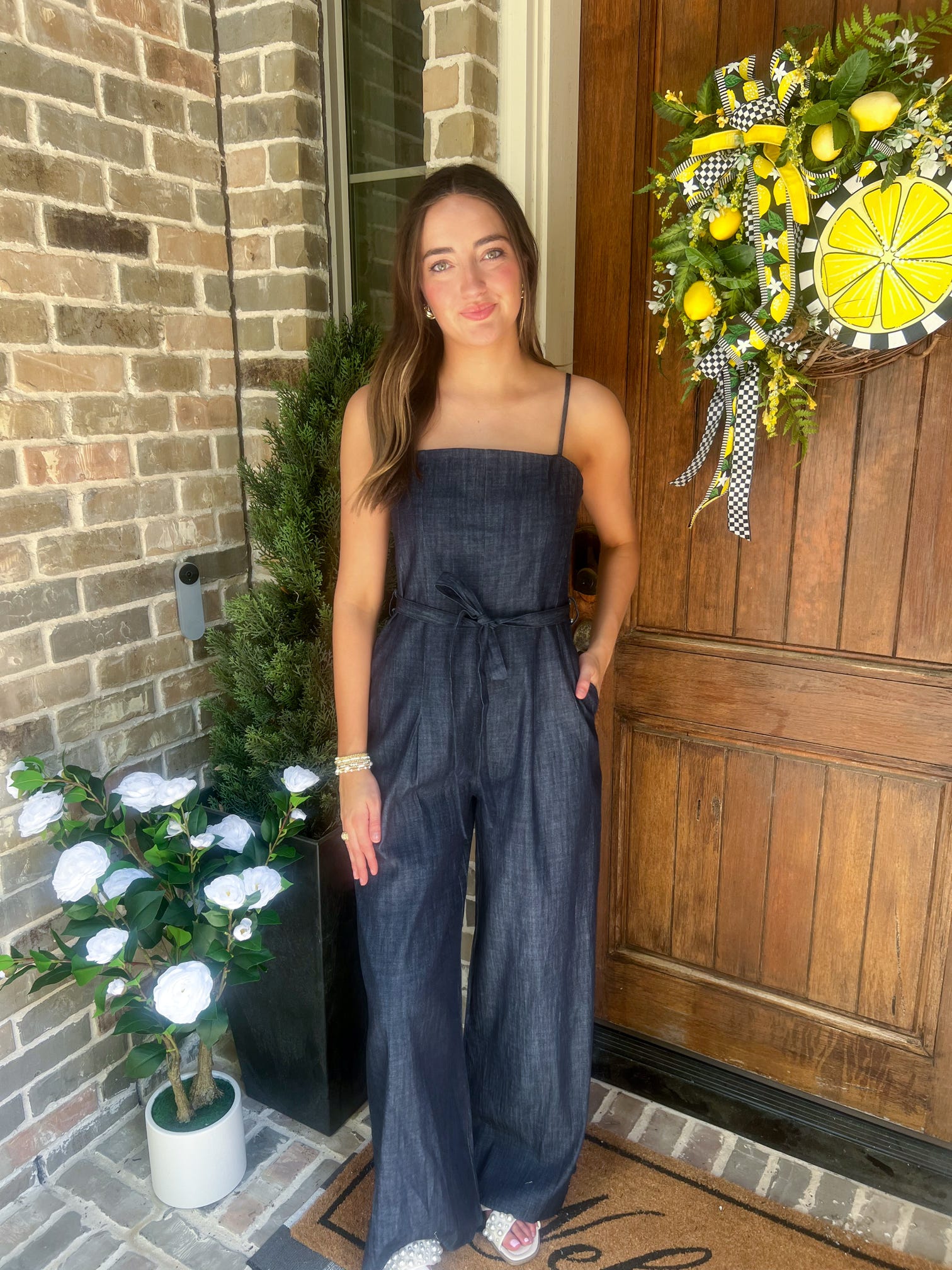 Woman in a denim jumpsuit standing in front of a wooden door with a decorative wreath.