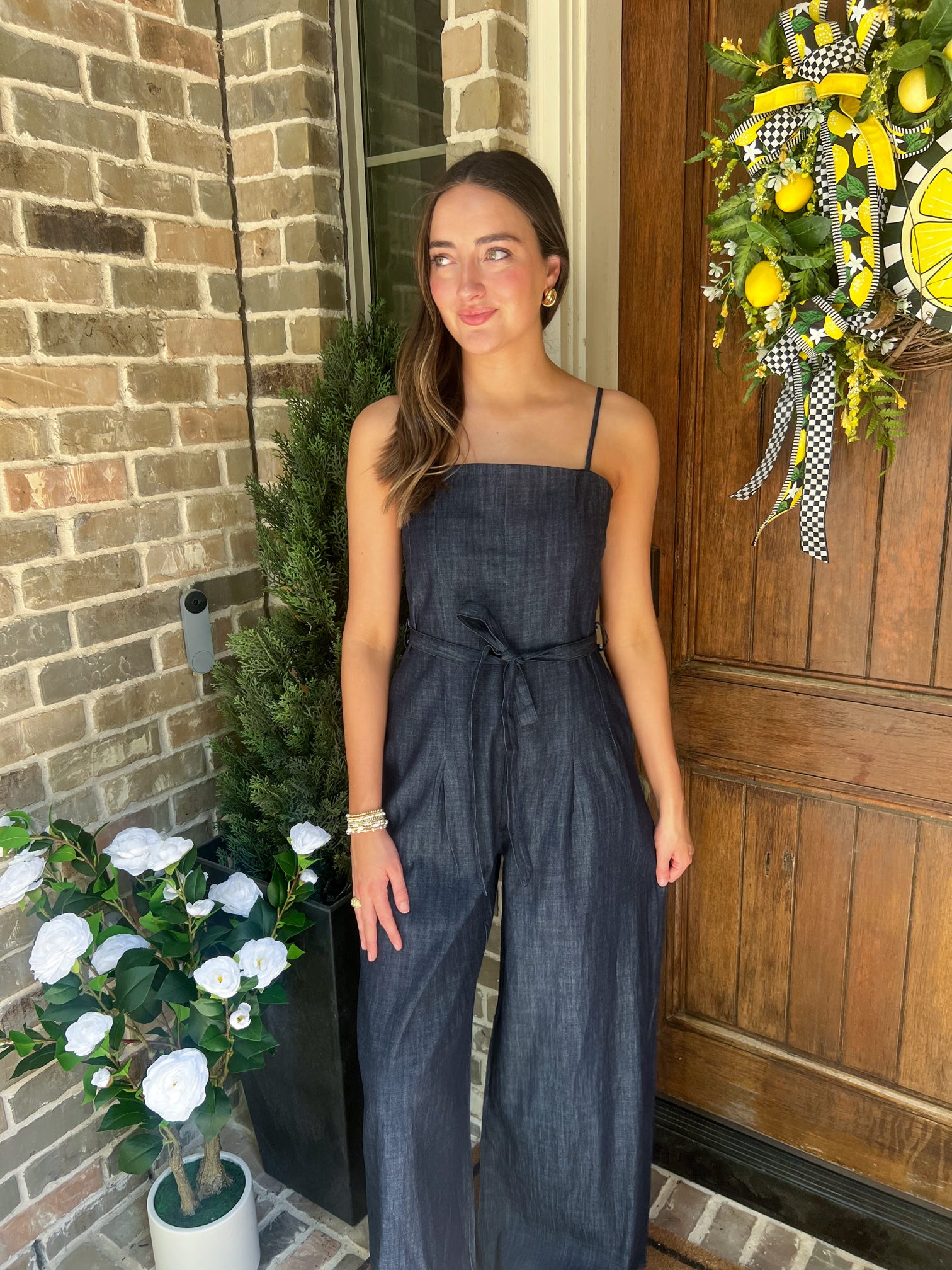 Woman in a denim jumpsuit standing on a porch with a wreath and plants.