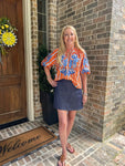 Model wearing an orange striped blouse with blue embroidered flowers standing in front of a wooden door