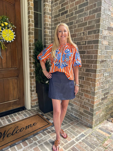 Model wearing an orange striped blouse with blue embroidered flowers standing in front of a wooden door