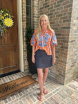 Model wearing an orange striped blouse with blue embroidered flowers standing in front of a wooden door