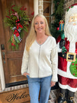 Woman wearing an off-white long sleeve v-neck sweater with a black line around the collar and sleeves and down the center of the sweater standing in front of a door with Christmas decorations, including a wreath and a Santa Claus figure.