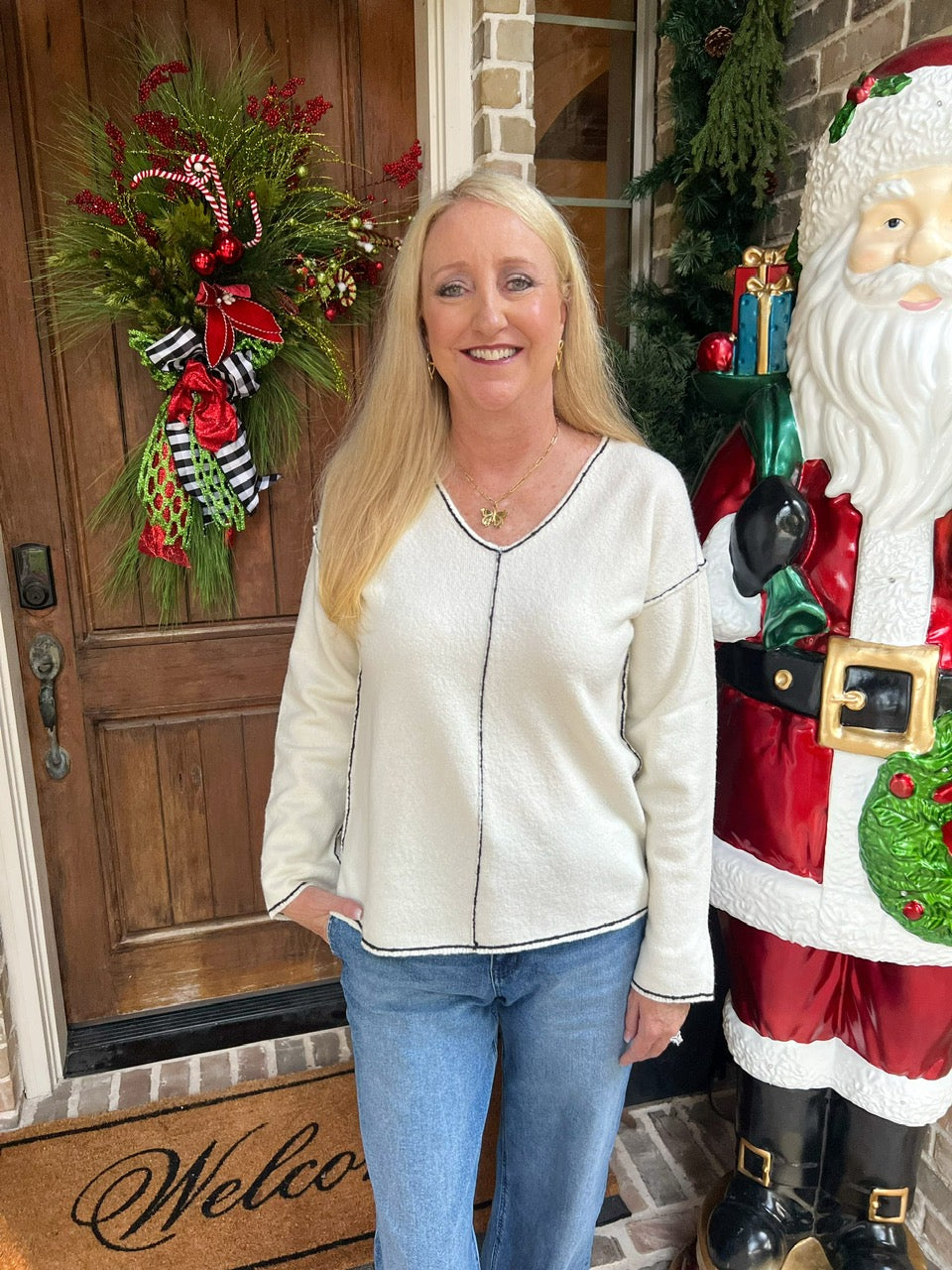 Woman wearing an off-white long sleeve v-neck sweater with a black line around the collar and sleeves and down the center of the sweater standing in front of a door with Christmas decorations, including a wreath and a Santa Claus figure.