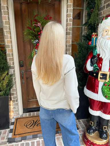 Woman modeling the back of an off-white long sleeve v-neck sweater with a black line around the collar and sleeves and down the center of the sweater standing in front of a door with Christmas decorations, including a wreath and a Santa Claus figure.