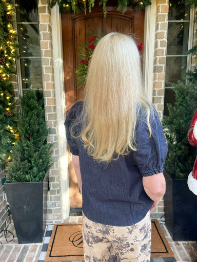 Woman modeling the back of a dark denim blouse with short bubble sleeves and a v-neck collar in front of a door with Christmas decorations.