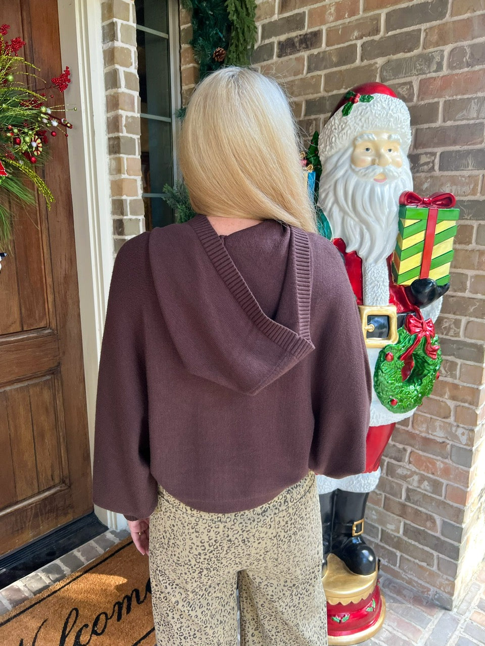 Woman modeling the back a chocolate brown long sleeve hooded sweater with tan cheetah print pants standing in front of a door with Christmas decorations
