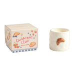 A small white candle shaped like a croissant with a butter scent sits beside its illustrated cream-colored box labeled 'Croissant Club' on a plain white background.