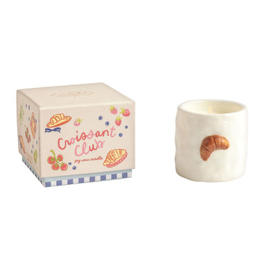 A small white candle shaped like a croissant with a butter scent sits beside its illustrated cream-colored box labeled 'Croissant Club' on a plain white background.