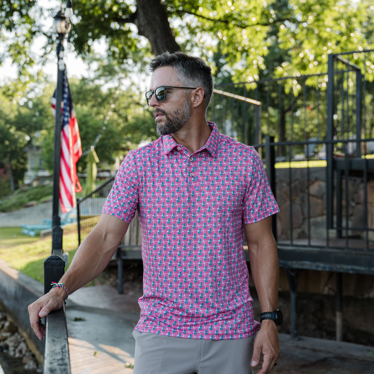 Man wearing an American Flag patterned short sleeve polo shirt standing outdoors with trees and an American flag in the background.