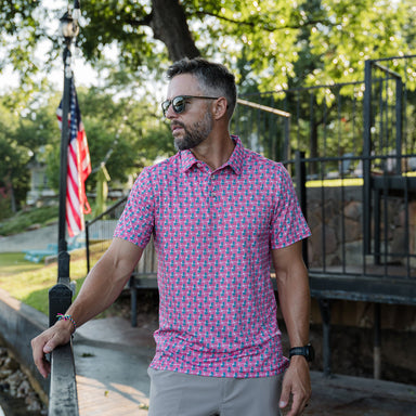 Man wearing an American Flag patterned short sleeve polo shirt standing outdoors with trees and an American flag in the background.
