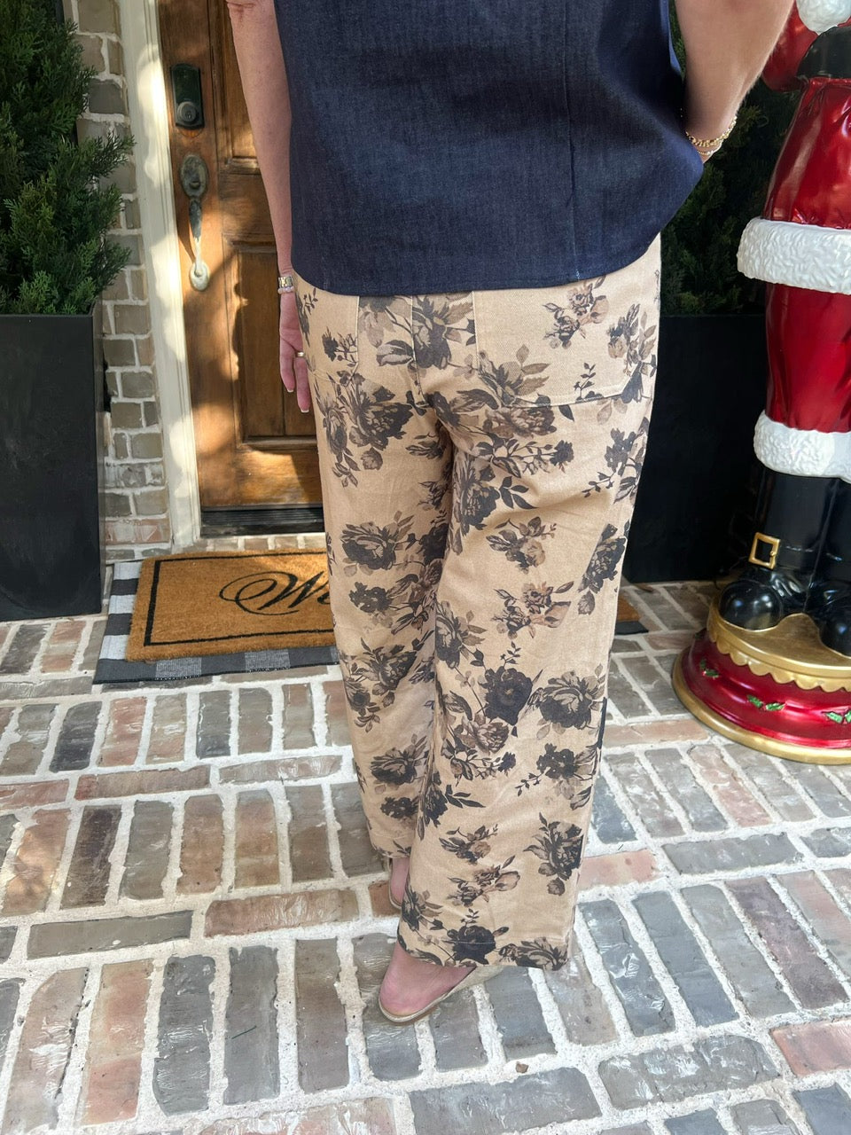 Woman modeling beige floral pants standing on a brick porch with a 'Welcome' mat.