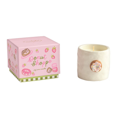 A donut scented candle in a textured cream-colored ceramic pot with a donut illustration on the front, shown next to its pink gift box decorated with donuts, strawberries, and macarons. The brand 'Candle Shoppe' is visible on the packaging.