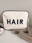 Photo of an XL light beige canvas accessory or makeup bag with a black trim and black patch letters saying Hair sewn on the front 