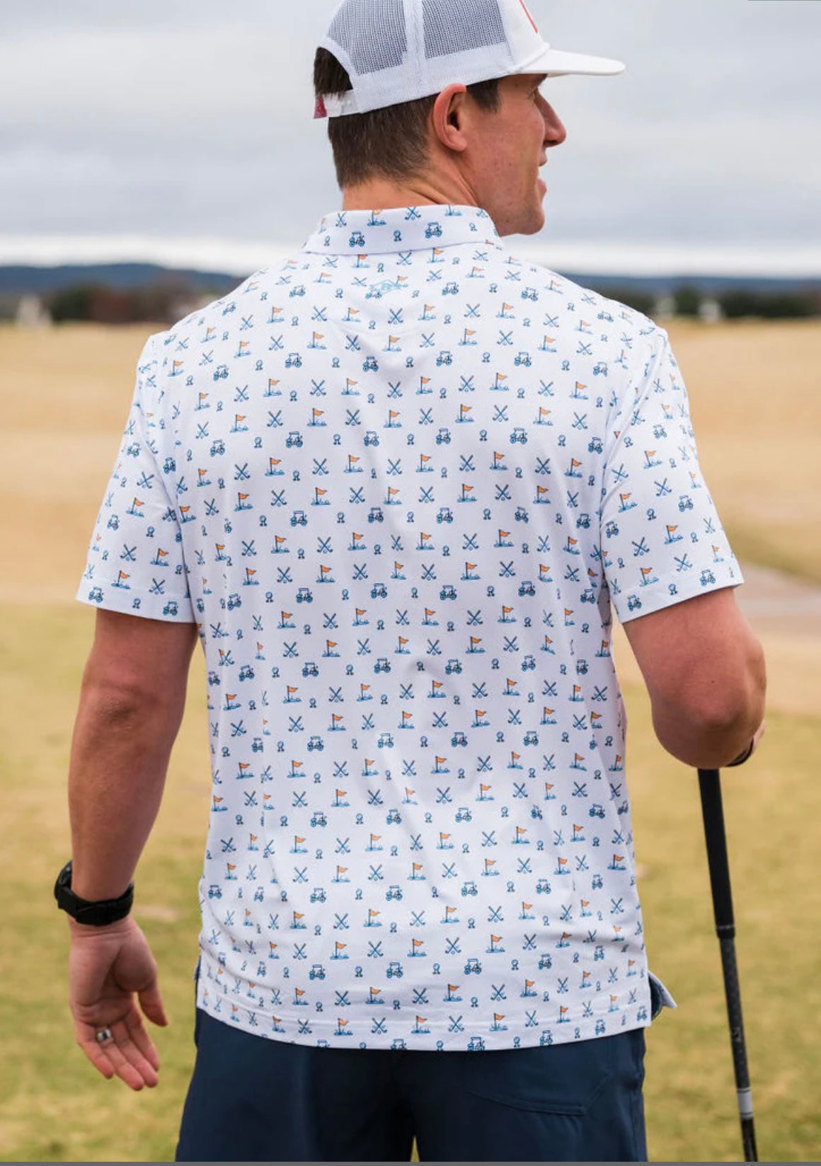 Photo of a man modeling the back of our performance white short sleeve polo featuring a fun print of a golf ball, cart, and putting green in shades of blue.