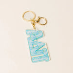 A Greek letter keychain featuring the letters 'ΑDPi' in light blue with a floral pattern, attached to a gold-tone lobster clasp and split ring, set against a light cream background.