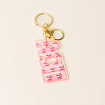 A pink acrylic keychain featuring the word 'AOPi' in stylized letters filled with pink bows and small floral details, attached to gold-tone metal rings on a light beige background.