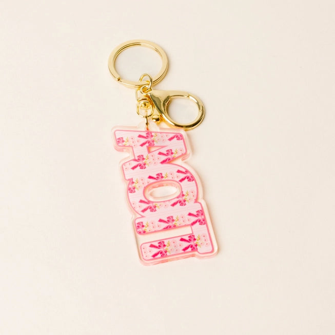 A pink acrylic keychain featuring the word 'AOPi' in stylized letters filled with pink bows and small floral details, attached to gold-tone metal rings on a light beige background.