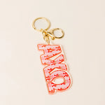 A pink acrylic keychain featuring the word 'AXO' in stylized letters filled with pink bows and small floral details, attached to gold-tone metal rings on a light beige background.