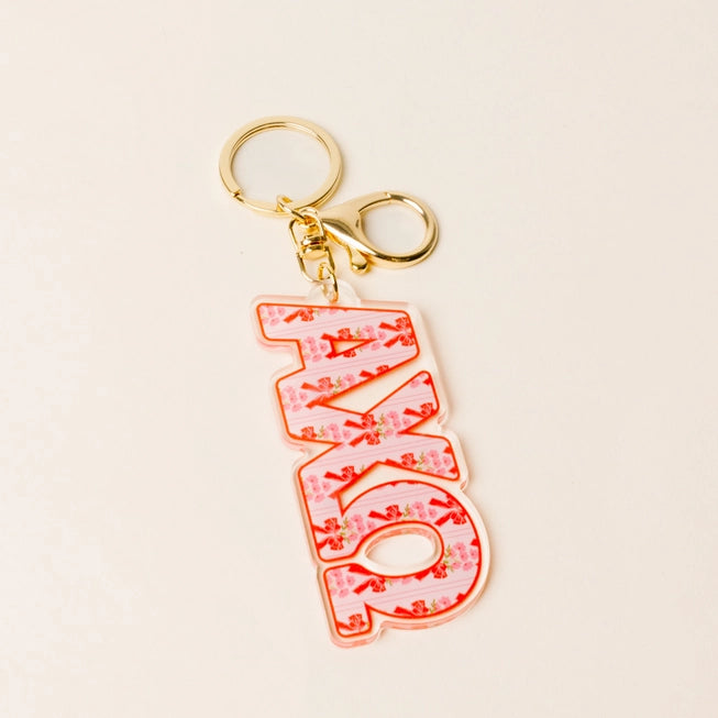 A pink acrylic keychain featuring the word 'AXO' in stylized letters filled with pink bows and small floral details, attached to gold-tone metal rings on a light beige background.