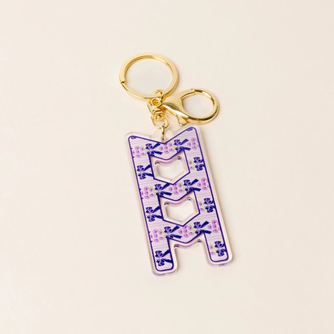 A pink acrylic keychain featuring the Greek letters for Sigma Sigma Sigma in stylized letters filled with pink bows and small floral details, attached to gold-tone metal rings on a light beige background.