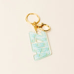A pink acrylic keychain featuring the word 'ZTA' in stylized letters filled with pink bows and small floral details, attached to gold-tone metal rings on a light beige background.