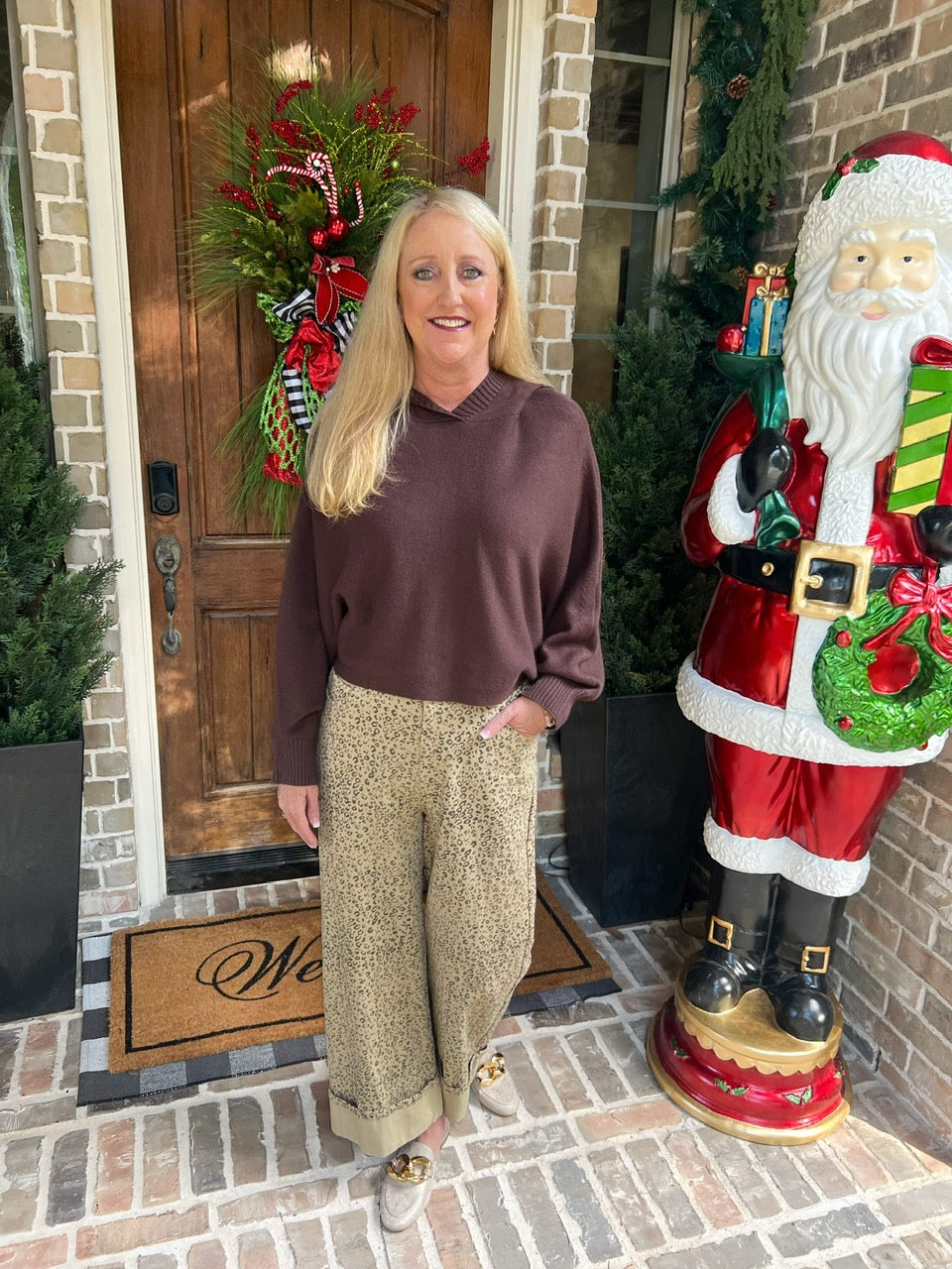 A woman wearing leopard print pants with a chocolate brown sweater standing on a brick sidewalk with a wooden door and doormat in the background.