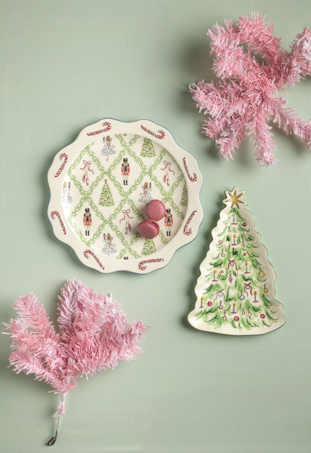A styled flat-lay featuring a white ceramic Christmas plate with a nutcracker and tree pattern, alongside a matching Christmas tree-shaped dish, pink decorative garlands, and two pink candies on a sage green background.