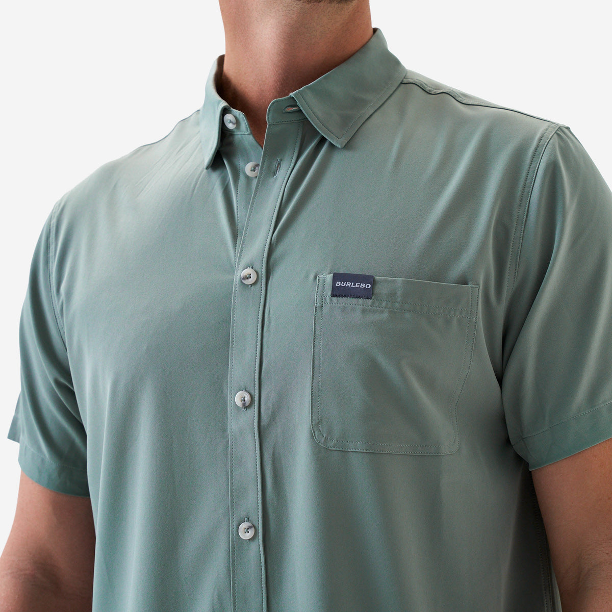 Up close photo of a sage green button-up shirt with a brand logo on a white background