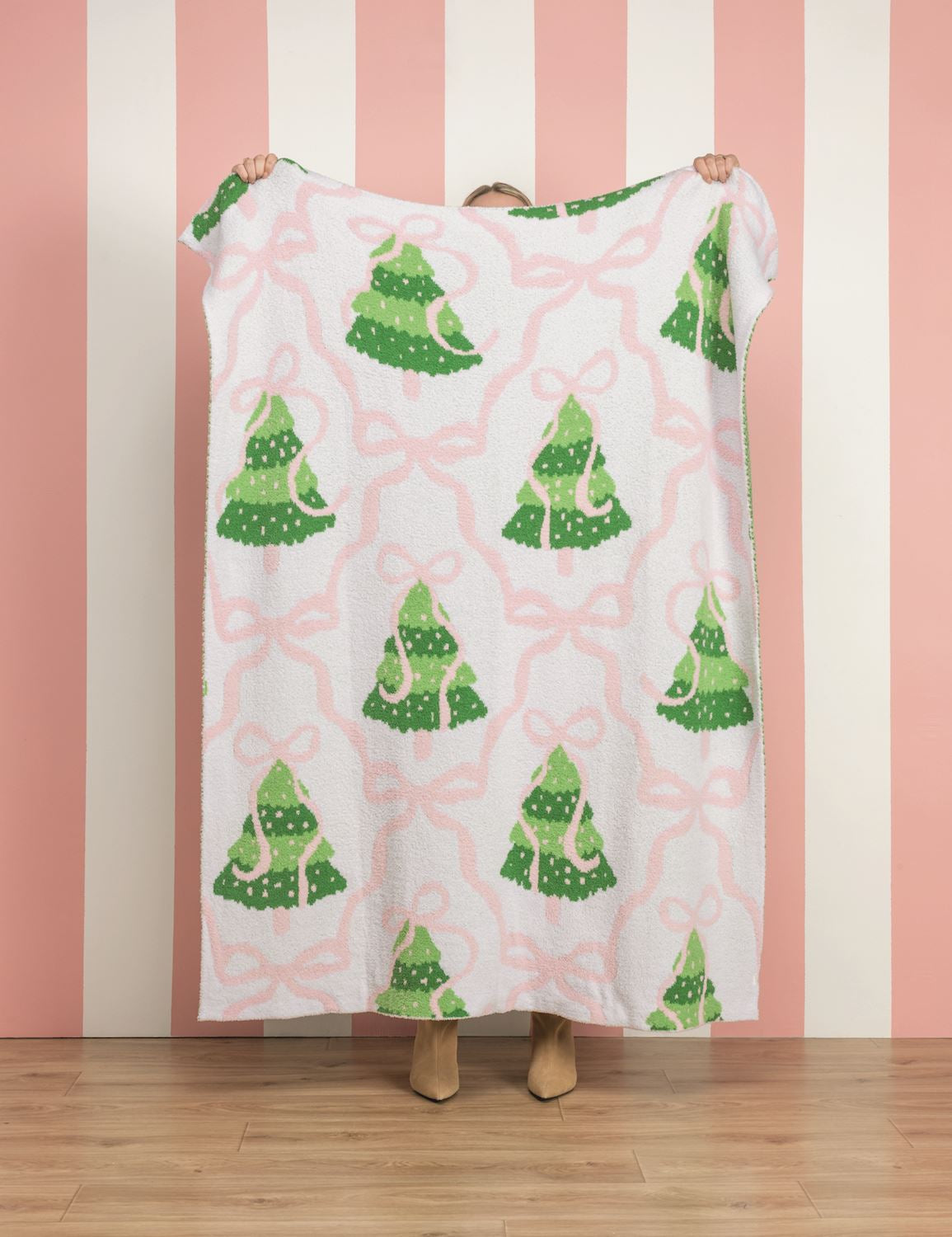 A person holds up a white fleece blanket featuring a green Christmas tree and pink bow pattern, set against a pink and white striped wall and wooden floor.