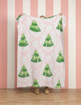 A person holds up a white fleece blanket featuring a green Christmas tree and pink bow pattern, set against a pink and white striped wall and wooden floor.