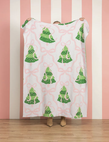 A person holds up a white fleece blanket featuring a green Christmas tree and pink bow pattern, set against a pink and white striped wall and wooden floor.