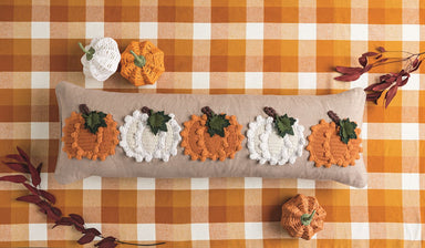 A long rectangular beige pillow with five crocheted pumpkins in orange and white, each with green leaves, displayed on an orange and white checkered blanket with autumn leaves and yarn balls.