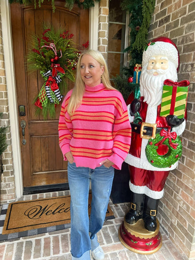 Woman in a pink sweater with orange stripes standing next to a large Santa Claus figure on a porch with Christmas decorations.