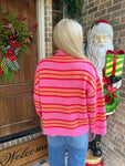 Person modeling a pink sweater with orange and red stripes standing next to a Santa Claus decoration on a porch.