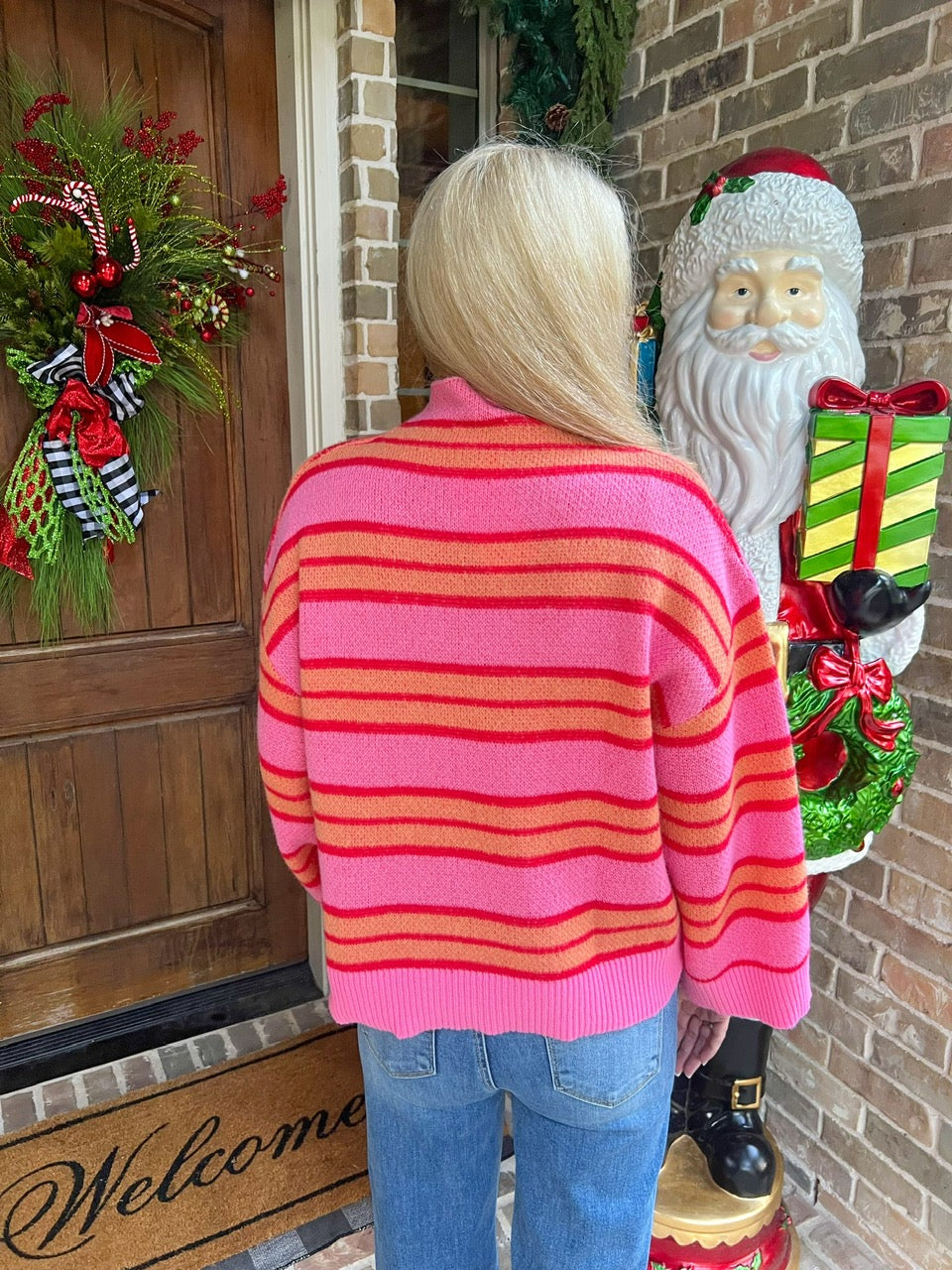 Person modeling a pink sweater with orange and red stripes standing next to a Santa Claus decoration on a porch.