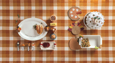 A display of Thanksgiving-inspired serving dishes, spreaders and potmitts on an orange and white checkered tablecloth.