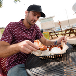  Man wearing a maroon short sleeve polo with white pennants saying Howdy sitting at a table outside eating food