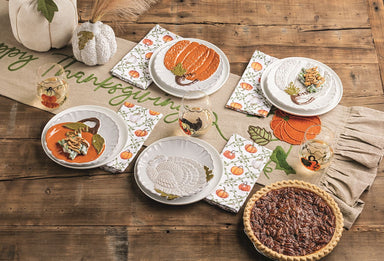 A styled Thanksgiving table setting with white plates, patterned napkins featuring pumpkins and turkeys, small glass candle holders, a burlap runner with green script, and a whole pecan pie on a rustic wooden table.