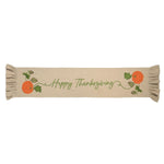 A beige fabric table runner with 'Happy Thanksgiving' embroidered in green script, decorated with orange pumpkins, green leaves, and pinecones on both ends, finished with ruffled edges.