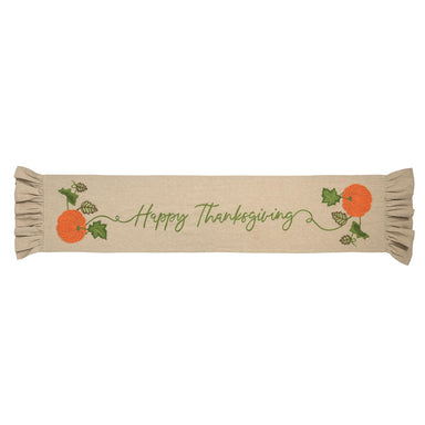 A beige fabric table runner with 'Happy Thanksgiving' embroidered in green script, decorated with orange pumpkins, green leaves, and pinecones on both ends, finished with ruffled edges.