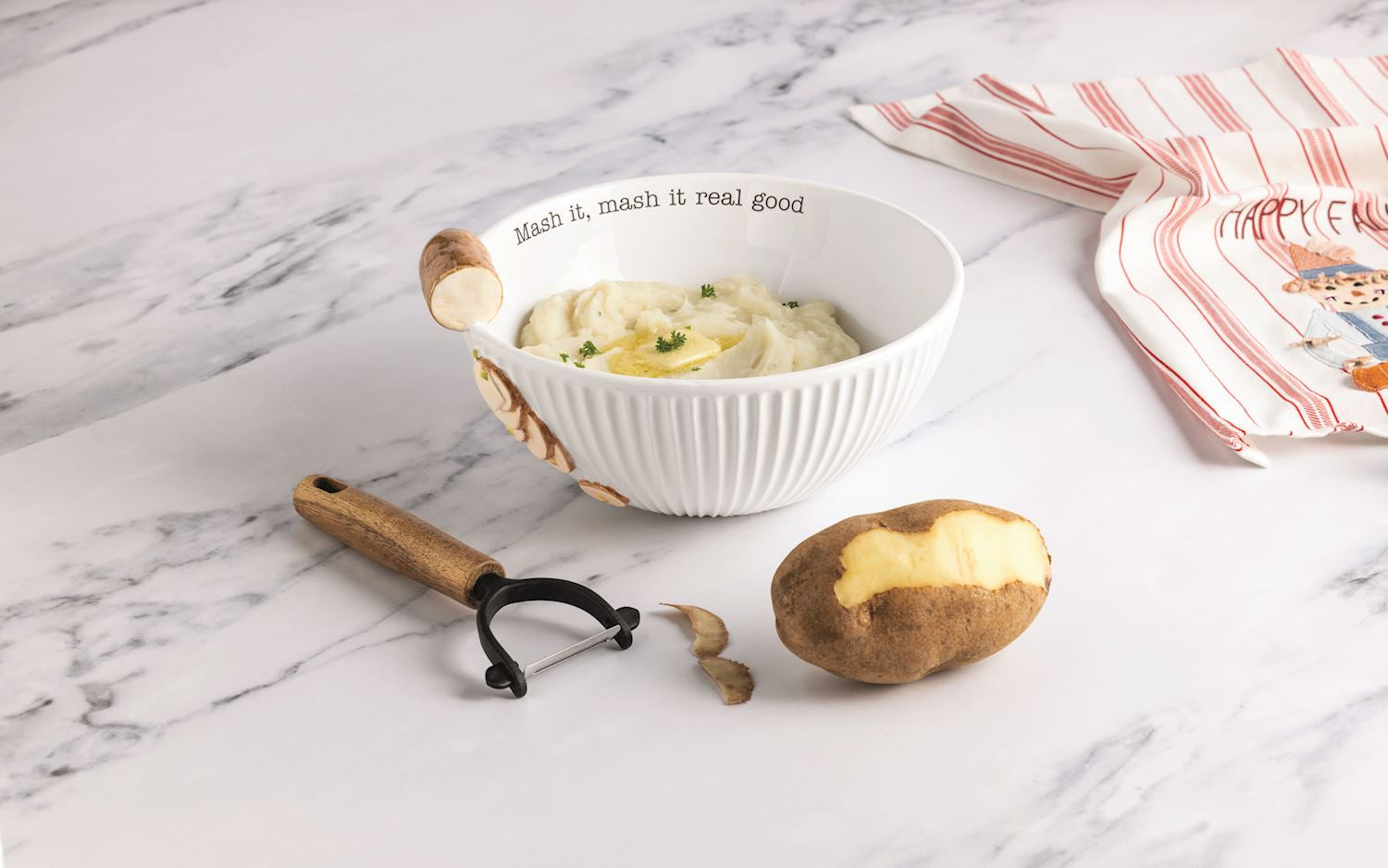 A white ribbed ceramic bowl with a wooden spoon handle and printed text sits on a white marble surface, filled with mashed potatoes. A potato peeler lies beside it, with a partially peeled potato and a striped cloth napkin nearby.