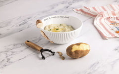 A white ribbed ceramic bowl with a wooden spoon handle and printed text sits on a white marble surface, filled with mashed potatoes. A potato peeler lies beside it, with a partially peeled potato and a striped cloth napkin nearby.