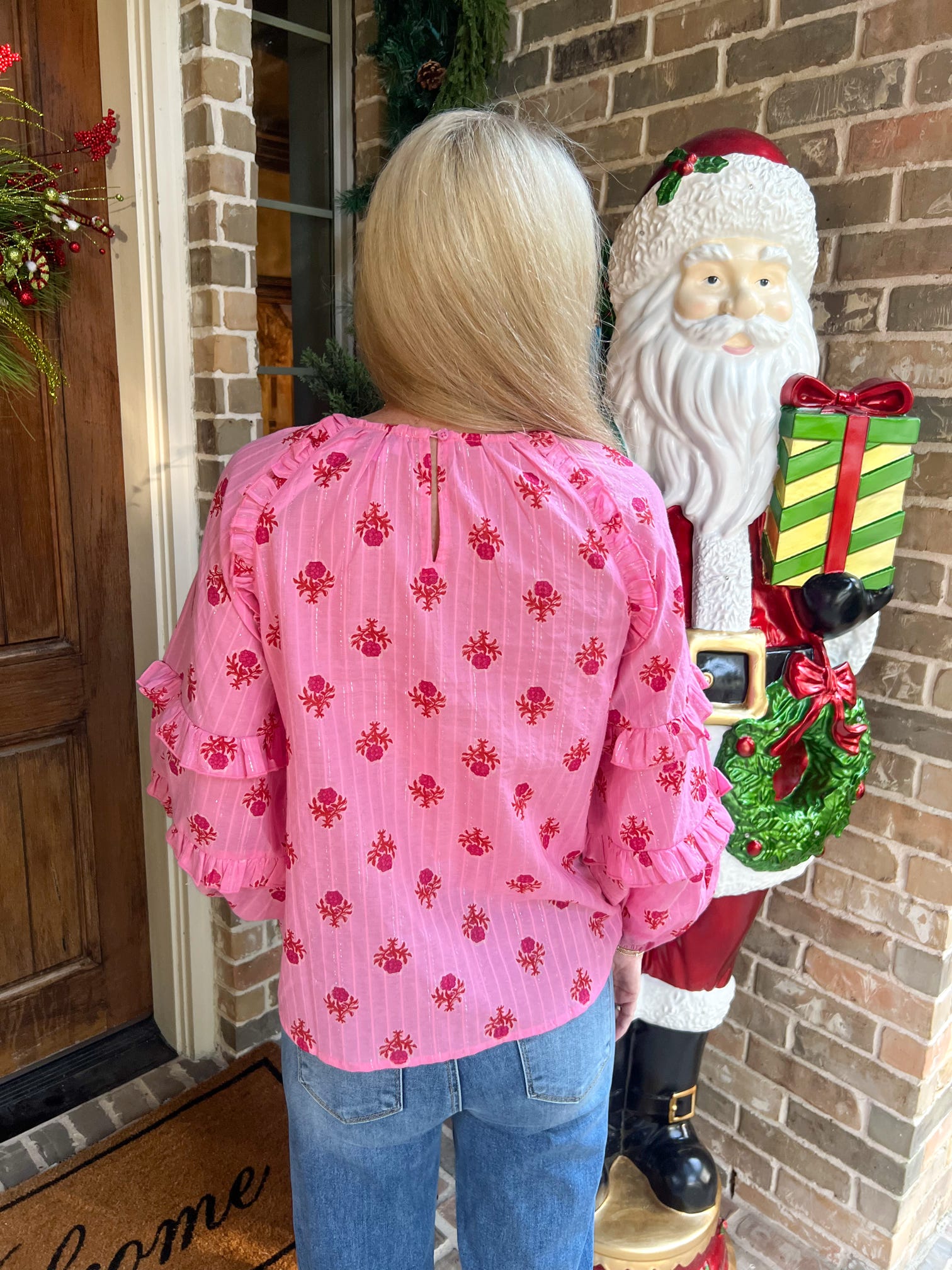 Person modeling the back of a pink floral blouse standing next to a Santa Claus statue with a wreath and gift.