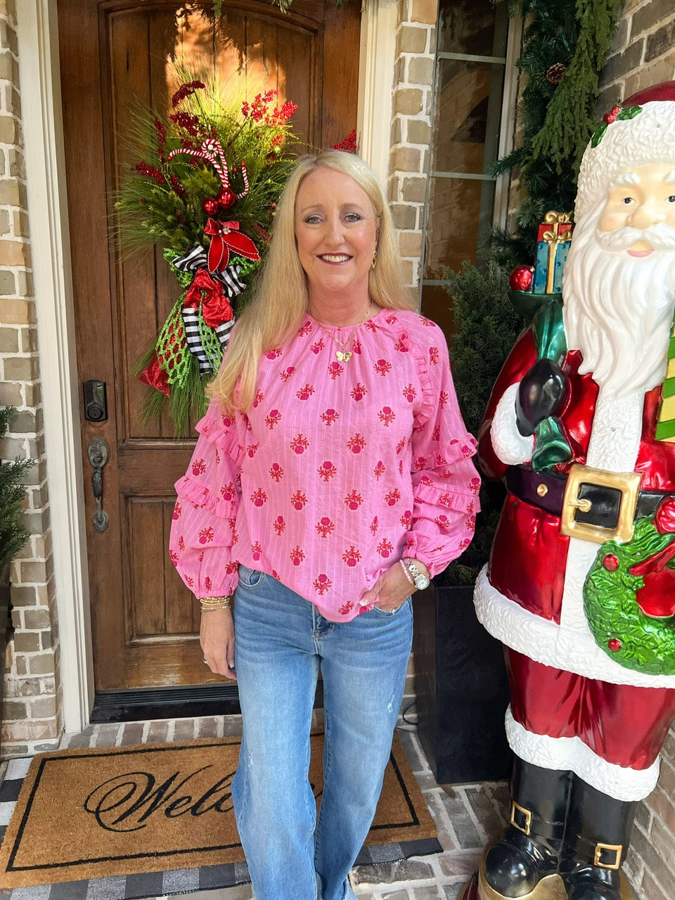 Woman in pink long sleeve blouse with red small flowers all over and jeans standing next to a Santa Claus statue on a porch with Christmas decorations.