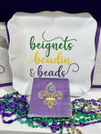 Photo of a white square serving platter with the words "Beignets, Boudin & Beads" written on the front in the traditional Mardi Gras colors